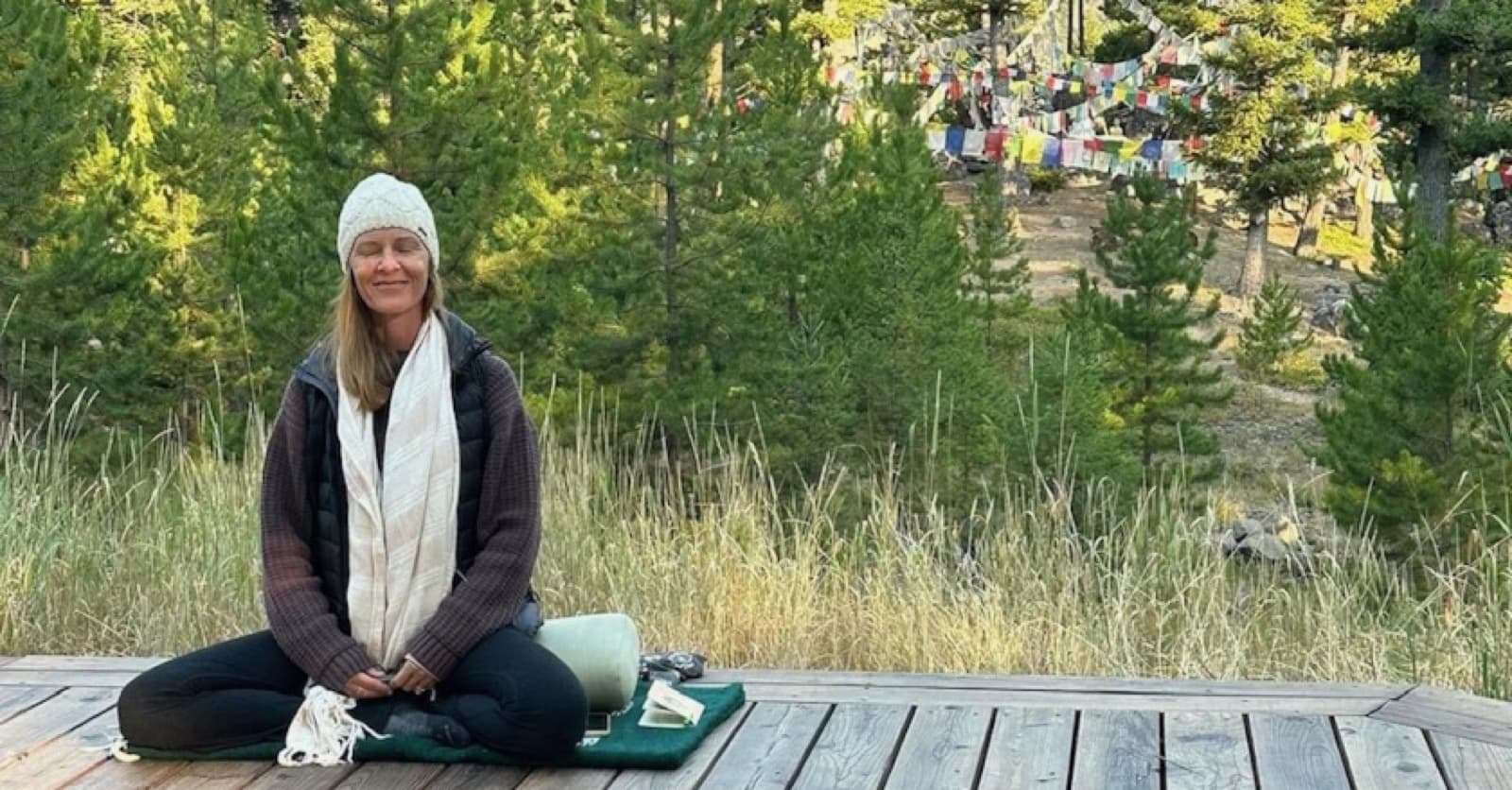 Jennifer French seated in meditation outdoors at Feathered Pipe Ranch with pine trees and prayer flags behind her